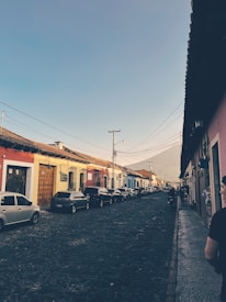 A picturesque cobblestone street lined with parked cars and colorful colonial-style buildings. The scene is set during the day with a clear sky, and a distant volcano is visible under the blue sky on the horizon. People can be seen walking along the street.