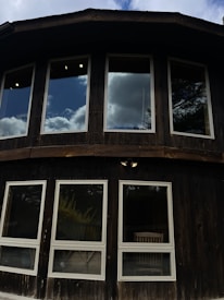 A wooden building facade featuring two rows of windows with reflections of clouds and sky, creating an impression of openness and connection with nature.