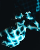 A person is illuminated by a blue and white grid pattern projected onto their body and face. The person is wearing dark sunglasses and a white sleeveless top, and their hand is adjusting the sunglasses. The background is dark, enhancing the contrast with the bright grid pattern.
