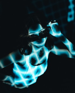 A person is illuminated by a blue and white grid pattern projected onto their body and face. The person is wearing dark sunglasses and a white sleeveless top, and their hand is adjusting the sunglasses. The background is dark, enhancing the contrast with the bright grid pattern.