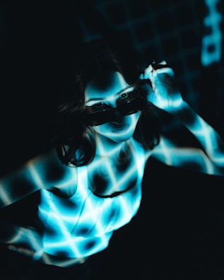 A person is illuminated by a blue and white grid pattern projected onto their body and face. The person is wearing dark sunglasses and a white sleeveless top, and their hand is adjusting the sunglasses. The background is dark, enhancing the contrast with the bright grid pattern.