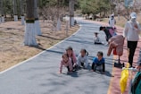 A group of young children are crawling on a paved path in a park setting. The path is bordered by trees with white painted trunks. Nearby, some adults are engaging with the children, with one person kneeling down and another standing with a scooter and an inflatable object.