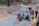 A group of young children are crawling on a paved path in a park setting. The path is bordered by trees with white painted trunks. Nearby, some adults are engaging with the children, with one person kneeling down and another standing with a scooter and an inflatable object.