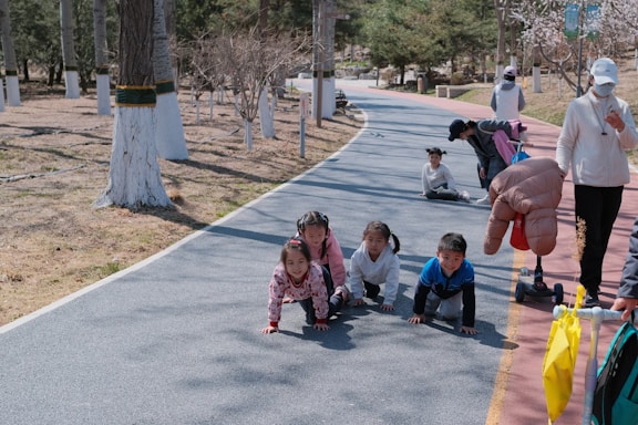 A group of young children are crawling on a paved path in a park setting. The path is bordered by trees with white painted trunks. Nearby, some adults are engaging with the children, with one person kneeling down and another standing with a scooter and an inflatable object.