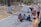 A group of young children are crawling on a paved path in a park setting. The path is bordered by trees with white painted trunks. Nearby, some adults are engaging with the children, with one person kneeling down and another standing with a scooter and an inflatable object.