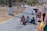 A group of young children are crawling on a paved path in a park setting. The path is bordered by trees with white painted trunks. Nearby, some adults are engaging with the children, with one person kneeling down and another standing with a scooter and an inflatable object.