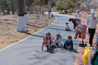 A group of young children are crawling on a paved path in a park setting. The path is bordered by trees with white painted trunks. Nearby, some adults are engaging with the children, with one person kneeling down and another standing with a scooter and an inflatable object.