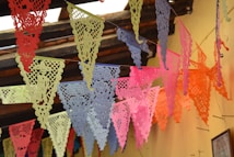 Brightly colored paper banners hang from the ceiling, featuring intricate cut-out designs. The banners are arranged in a festive, overlapping pattern against a rustic wooden ceiling and a light yellow wall.