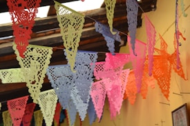 Brightly colored paper banners hang from the ceiling, featuring intricate cut-out designs. The banners are arranged in a festive, overlapping pattern against a rustic wooden ceiling and a light yellow wall.