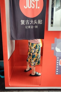 A person stands inside a photo booth with a curtain partially covering their upper body. The booth is bright red and there is text in a foreign language on the curtain. The person is wearing a colorful floral skirt and black slide sandals.