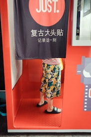 A person stands inside a photo booth with a curtain partially covering their upper body. The booth is bright red and there is text in a foreign language on the curtain. The person is wearing a colorful floral skirt and black slide sandals.