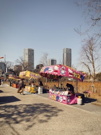 A row of colorful market stalls featuring printed tents with cartoon characters on them. People are walking and browsing the stalls, some of which are selling snacks or goods. Leafless trees are in the background along with tall modern skyscrapers against a clear sky.