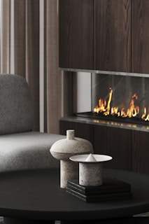 A modern living area featuring a sleek dark wood fireplace with visible flames. In the foreground, a black circular coffee table holds two stone-like sculptures with geometric designs. A plush gray chair is partially visible on the left, and vertically striped curtains add texture to the space.
