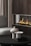 A modern living area featuring a sleek dark wood fireplace with visible flames. In the foreground, a black circular coffee table holds two stone-like sculptures with geometric designs. A plush gray chair is partially visible on the left, and vertically striped curtains add texture to the space.