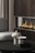 A modern living area featuring a sleek dark wood fireplace with visible flames. In the foreground, a black circular coffee table holds two stone-like sculptures with geometric designs. A plush gray chair is partially visible on the left, and vertically striped curtains add texture to the space.