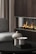 A modern living area featuring a sleek dark wood fireplace with visible flames. In the foreground, a black circular coffee table holds two stone-like sculptures with geometric designs. A plush gray chair is partially visible on the left, and vertically striped curtains add texture to the space.