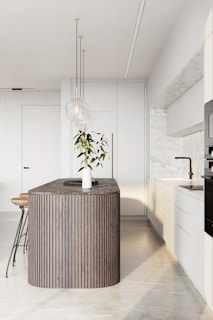 A modern kitchen featuring a sleek, minimalist design with a large, dark wood island in the center. The island is adorned with a decorative plant in a white vase. Three pendant lights hang above the island, providing elegant lighting. Surrounding the island are white cabinets with a marble backsplash, and bar stools with wooden seats and metal legs.