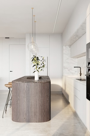 A modern kitchen featuring a sleek, minimalist design with a large, dark wood island in the center. The island is adorned with a decorative plant in a white vase. Three pendant lights hang above the island, providing elegant lighting. Surrounding the island are white cabinets with a marble backsplash, and bar stools with wooden seats and metal legs.