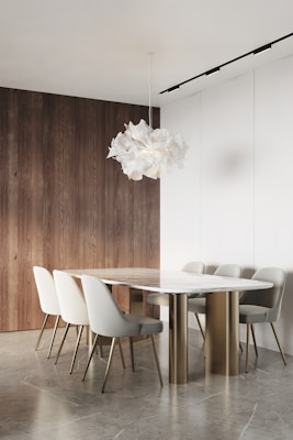 A minimalist dining room features a sleek marble table with a gold base surrounded by six modern upholstered chairs. The space is accented by a unique, sculptural white pendant light. The backdrop includes a combination of natural wood paneling and white walls, complementing the neutral tones of the floor.