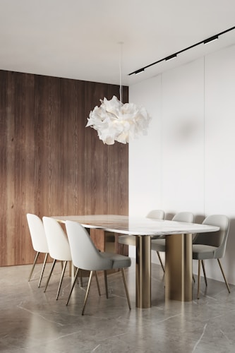 A minimalist dining room features a sleek marble table with a gold base surrounded by six modern upholstered chairs. The space is accented by a unique, sculptural white pendant light. The backdrop includes a combination of natural wood paneling and white walls, complementing the neutral tones of the floor.