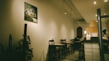 A cozy, dimly lit cafe interior featuring several small tables and chairs arranged along the wall. The decor includes a painting of flowers on the wall and some indoor plants adding greenery to the space. The floor is tiled in a retro style, and the counter area displays various items, suggesting a welcoming and intimate atmosphere.