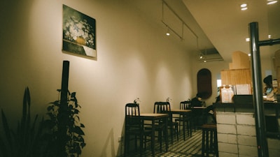 A cozy, dimly lit cafe interior featuring several small tables and chairs arranged along the wall. The decor includes a painting of flowers on the wall and some indoor plants adding greenery to the space. The floor is tiled in a retro style, and the counter area displays various items, suggesting a welcoming and intimate atmosphere.