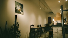 A cozy, dimly lit cafe interior featuring several small tables and chairs arranged along the wall. The decor includes a painting of flowers on the wall and some indoor plants adding greenery to the space. The floor is tiled in a retro style, and the counter area displays various items, suggesting a welcoming and intimate atmosphere.