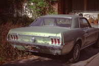 A vintage, pale green Ford Mustang parked in a driveway, surrounded by foliage. The car's classic taillights and chrome bumper are visible, along with a California license plate.
