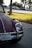 A classic car with a rounded shape is parked on the side of a road in a sunlit area with grass and trees in the background. It features a California license plate and has a maroon color with chrome accents.