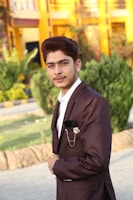 A young man in a dark suit with a decorative lapel pin stands outdoors, with a landscaped garden and a bright yellow building in the background.