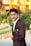 A young man in a dark suit with a decorative lapel pin stands outdoors, with a landscaped garden and a bright yellow building in the background.