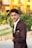 A young man in a dark suit with a decorative lapel pin stands outdoors, with a landscaped garden and a bright yellow building in the background.