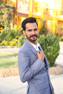 Portrait of Alcides Riveros smiling confidently in a blue suit, standing outdoors with a subtle Paraguayan flag in the background.