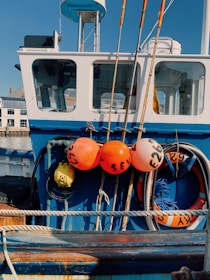 A close-up view of a fishing boat featuring several orange buoys with numbers written on them, various fishing equipment, and a life preserver. The cabin of the boat is visible with windows reflecting the surroundings. The boat is a bright blue color, with some ropes and pipes attached to its deck.