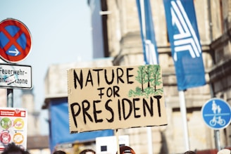 A protest sign with the words 'Nature for President' is held up in a street setting. The sign is decorated with green tree illustrations. Various street signs, including no parking and bike lane symbols, are visible. The background features urban architecture and blue flags with abstract designs.