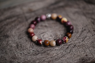 A bracelet made of polished beads in various shades of brown, red, and purple is placed on a textured wooden surface with a natural, rustic appearance. The beads are shiny and have a smooth finish, strung together in a circular arrangement.