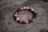 A bracelet made of polished beads in various shades of brown, red, and purple is placed on a textured wooden surface with a natural, rustic appearance. The beads are shiny and have a smooth finish, strung together in a circular arrangement.