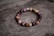 A bracelet made of polished beads in various shades of brown, red, and purple is placed on a textured wooden surface with a natural, rustic appearance. The beads are shiny and have a smooth finish, strung together in a circular arrangement.