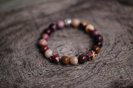 A bracelet made of polished beads in various shades of brown, red, and purple is placed on a textured wooden surface with a natural, rustic appearance. The beads are shiny and have a smooth finish, strung together in a circular arrangement.