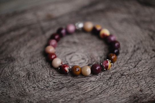 A bracelet made of polished beads in various shades of brown, red, and purple is placed on a textured wooden surface with a natural, rustic appearance. The beads are shiny and have a smooth finish, strung together in a circular arrangement.