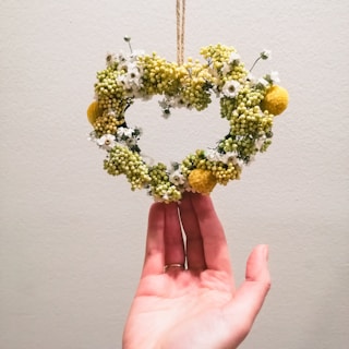 A hand is gently holding a heart-shaped wreath made of small yellow and white flowers. The wreath is suspended by a thin rope against a plain light-colored wall.
