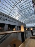 A spacious interior of a modern building featuring a high, geometric glass ceiling that lets in natural light. Below the ceiling are two stories, with visible escalators and railings. A sign with the words 'COMING SOON' is prominently displayed. Soft lighting on tall stands creates a warm and inviting atmosphere.