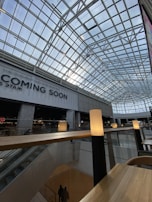 A spacious interior of a modern building featuring a high, geometric glass ceiling that lets in natural light. Below the ceiling are two stories, with visible escalators and railings. A sign with the words 'COMING SOON' is prominently displayed. Soft lighting on tall stands creates a warm and inviting atmosphere.