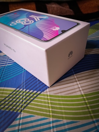 A Huawei Y9a smartphone is packaged in a sleek, white rectangular box placed on a colorful, patterned surface. The box features the Huawei logo and the smartphone image with vibrant shades of blue and pink displayed on the screen.