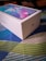 A Huawei Y9a smartphone is packaged in a sleek, white rectangular box placed on a colorful, patterned surface. The box features the Huawei logo and the smartphone image with vibrant shades of blue and pink displayed on the screen.