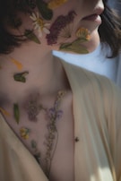 A close-up of a person with pressed flowers and leaves placed artfully on their face and neck, creating an artistic and natural look. The person is wearing a light yellow top and has soft lighting that creates gentle shadows.