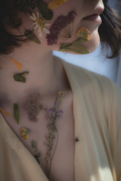A close-up of a person with pressed flowers and leaves placed artfully on their face and neck, creating an artistic and natural look. The person is wearing a light yellow top and has soft lighting that creates gentle shadows.