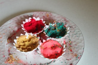 A white textured plate holds four small, scalloped bowls filled with vibrant colored powders in shades of pink, red, yellow, and teal. The surface of the plate and table is speckled with scattered powder, adding a splash of color.