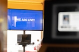 A large screen displays the message 'WE ARE LIVE!' in bold white lettering against a blue background. In front of the screen, there is a laptop on a stand showing an out-of-focus presentation slide. Another blurred screen is visible in the foreground, suggesting a busy digital or virtual environment.