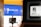 A large screen displays the message 'WE ARE LIVE!' in bold white lettering against a blue background. In front of the screen, there is a laptop on a stand showing an out-of-focus presentation slide. Another blurred screen is visible in the foreground, suggesting a busy digital or virtual environment.